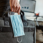 Person in an office holding a briefcase and a surgical mask in his hand.