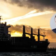 Silhouetted cargo ship at sea during a dramatic sunset, with dark clouds and golden light on the horizon; a circular inset shows a professional headshot of an economist.