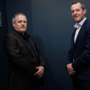 Two individuals standing in a dark blue office setting beside a wall-mounted FutureRange logo, dressed in formal business attire, facing the camera with hands clasped, in a professionally lit corporate portrait style.