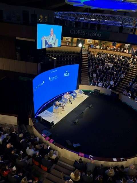 A large conference audience seated in a tiered indoor venue watches a speaker seated on stage. A curved digital screen displays event branding and sponsor logos, while a live video feed of the speaker appears on a screen above the seating area.