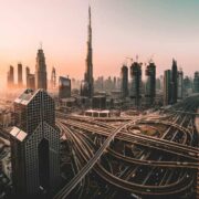 A wide aerial view of a modern Middle Eastern city skyline at sunset, featuring dense clusters of high-rise skyscrapers, including the Burj Khalifa at the centre, with multilayered highways and interchanges weaving through the foreground under warm golden light.